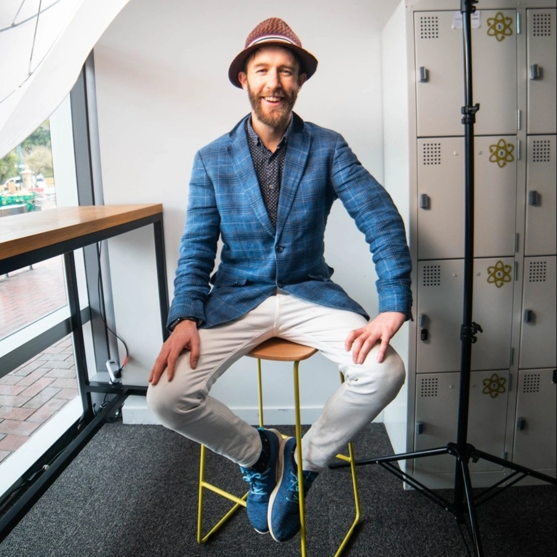 Logan Elliott in a hat and blue blazer seated on a stool by a window