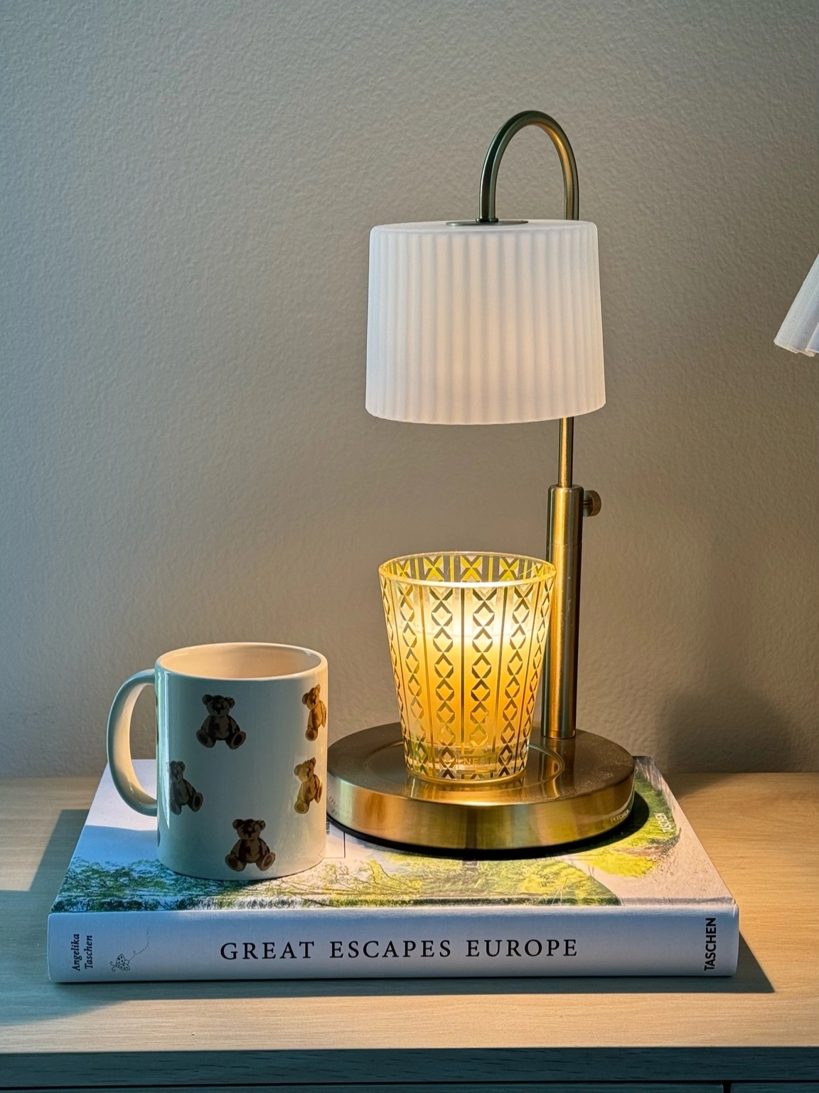 A candle warmer lamp glowing beside a teddy bear mug on a stack of books