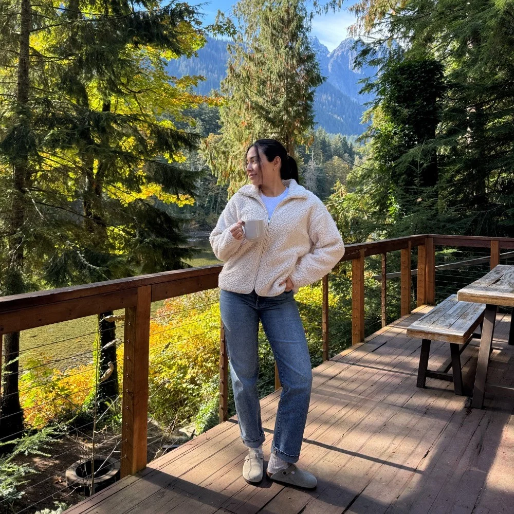 Arabella on the deck of a forest cabin with mountains behind her