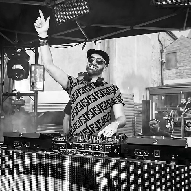 A DJ in a cap and sunglasses with one hand raised behind the decks at Darwen Live