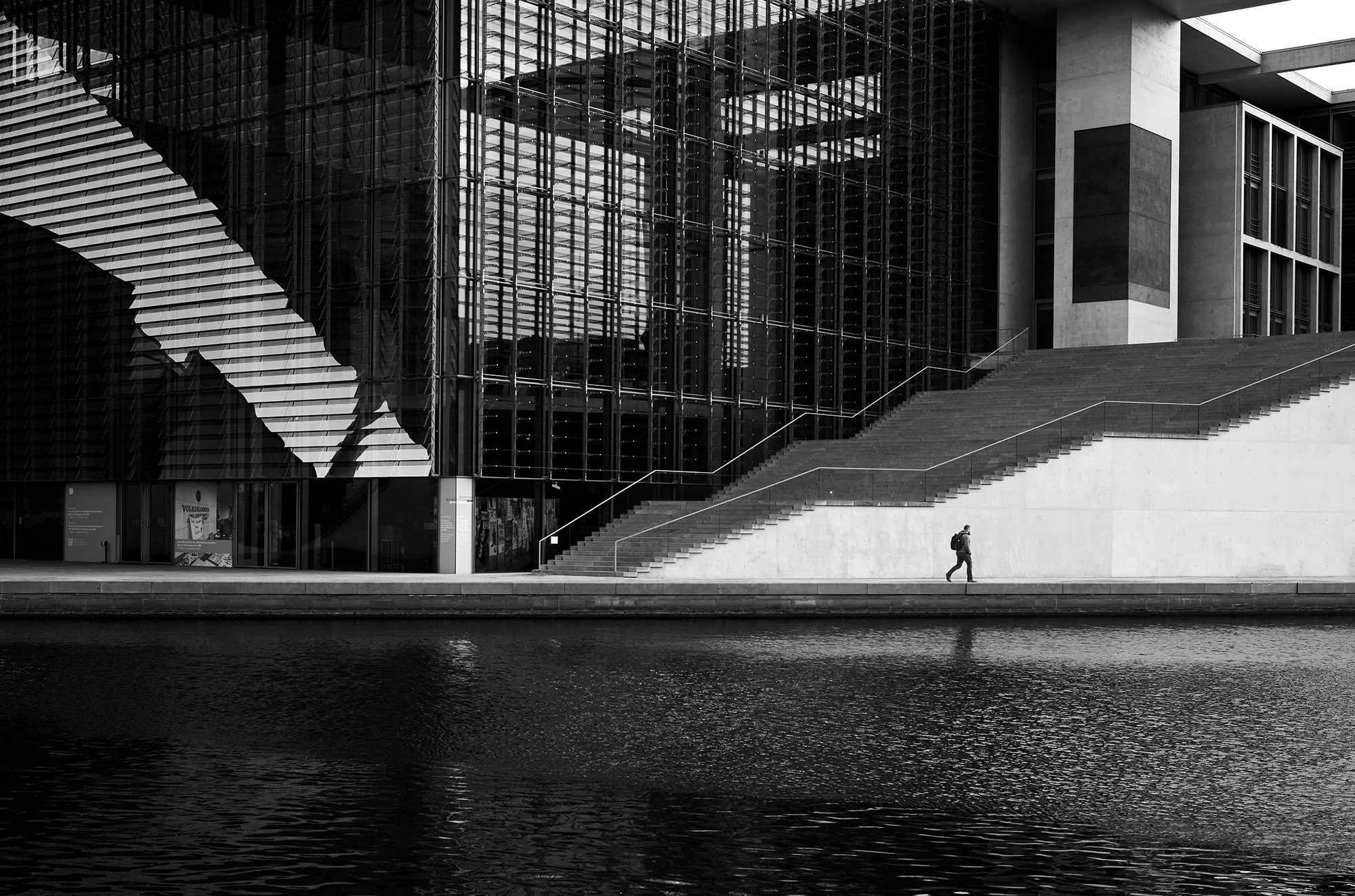 Una persona cammina lungo il fiume davanti a un grande edificio di vetro e cemento con una lunga scalinata