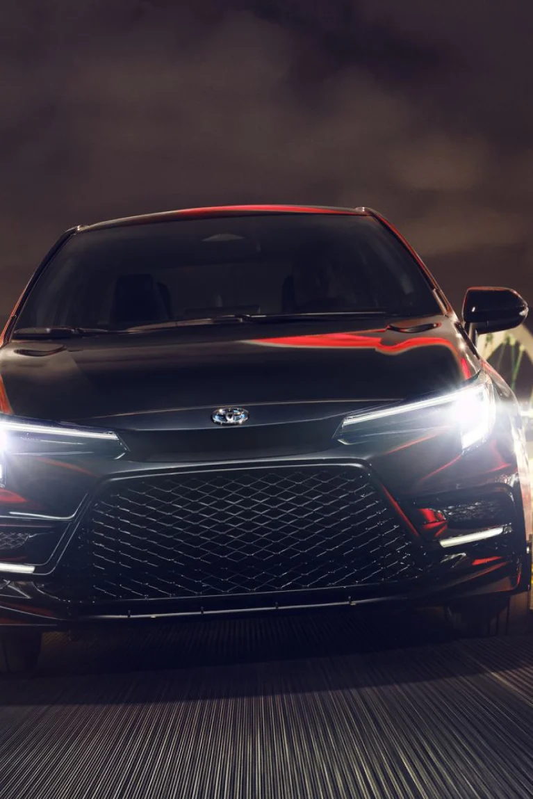 The front of a black Toyota Corolla with its headlights on
