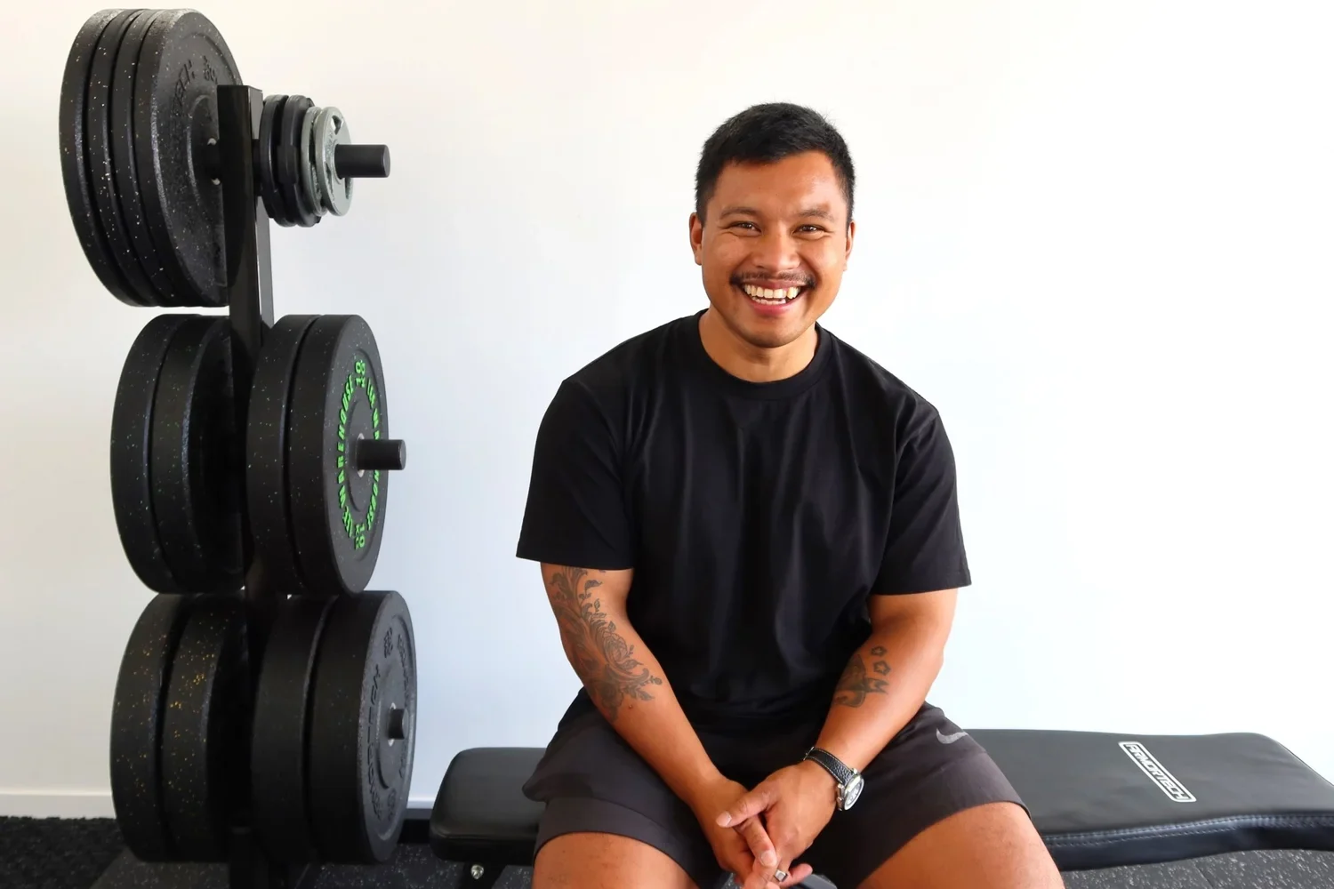 Marwin smiling beside a stack of weight plates in a bright training studio