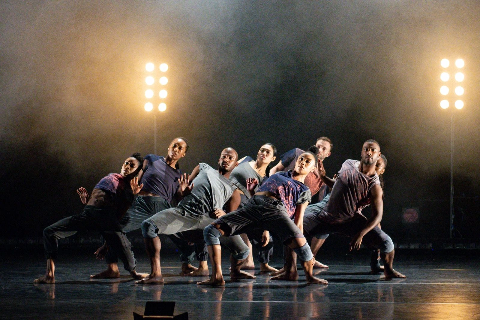The company in a synchronized low stance on a dark stage between two tall light rigs in Drive