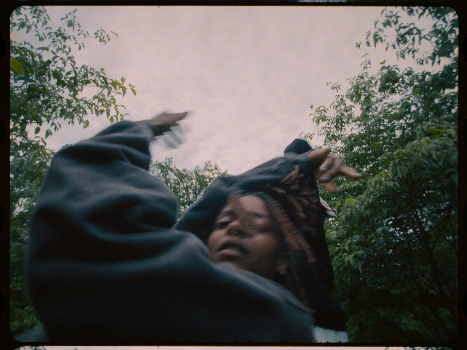 A dancer mid-gesture with an arm raised, framed by tree branches against an overcast sky