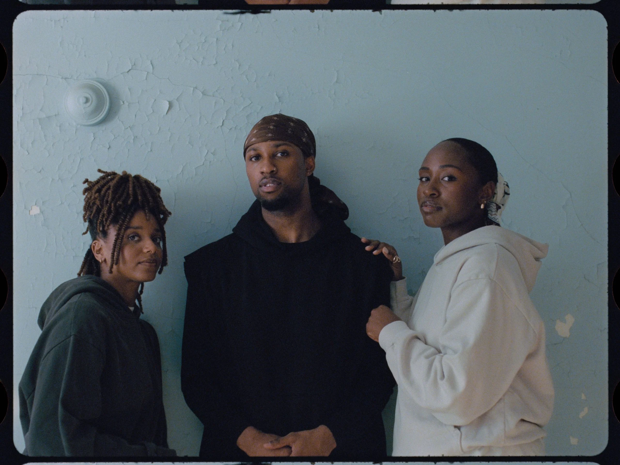 Three dancers stand shoulder to shoulder against a pale blue wall, looking at the camera
