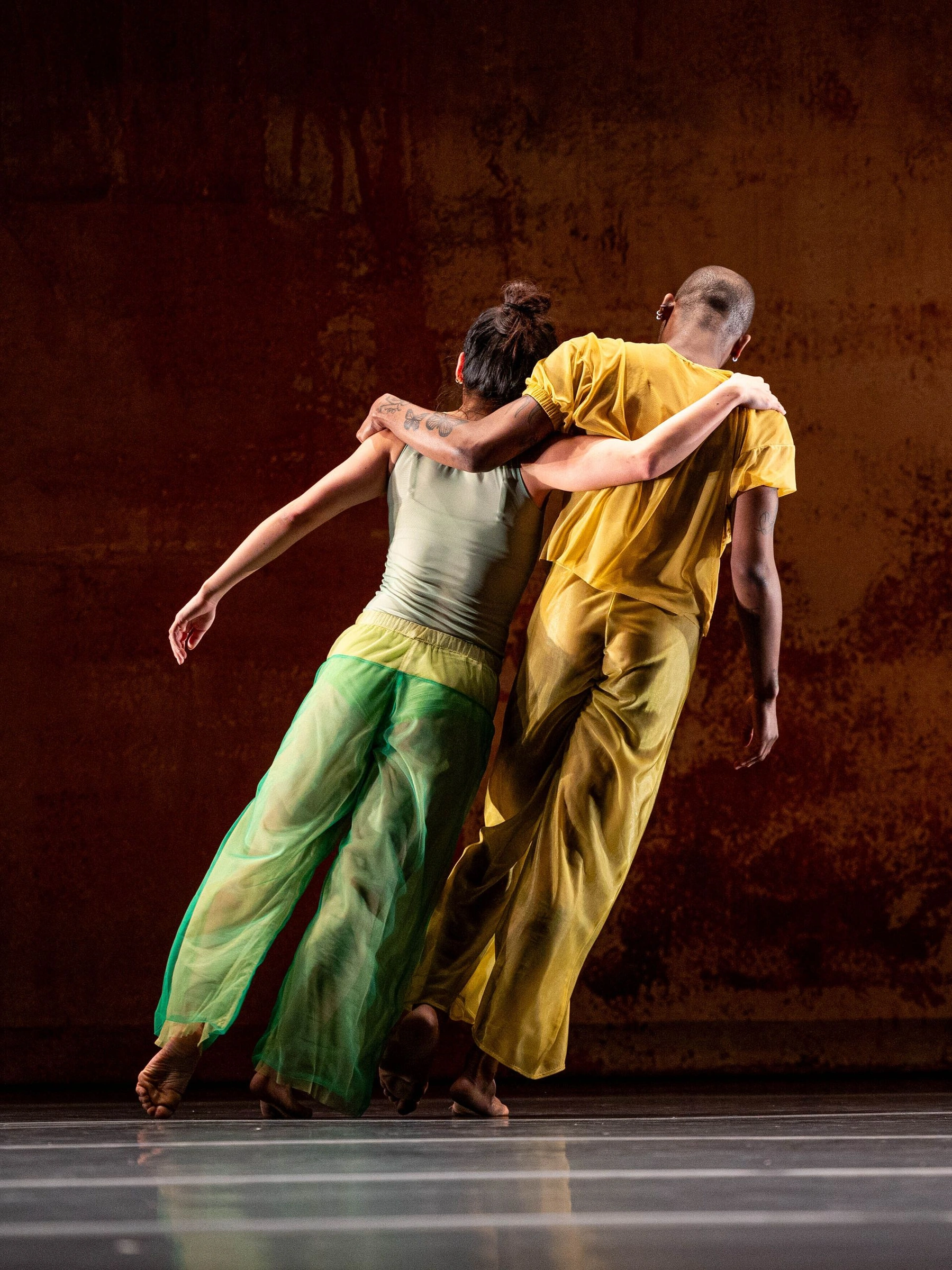 Two dancers in green and yellow lean on each other’s shoulders in front of a rust-colored backdrop in 2x4