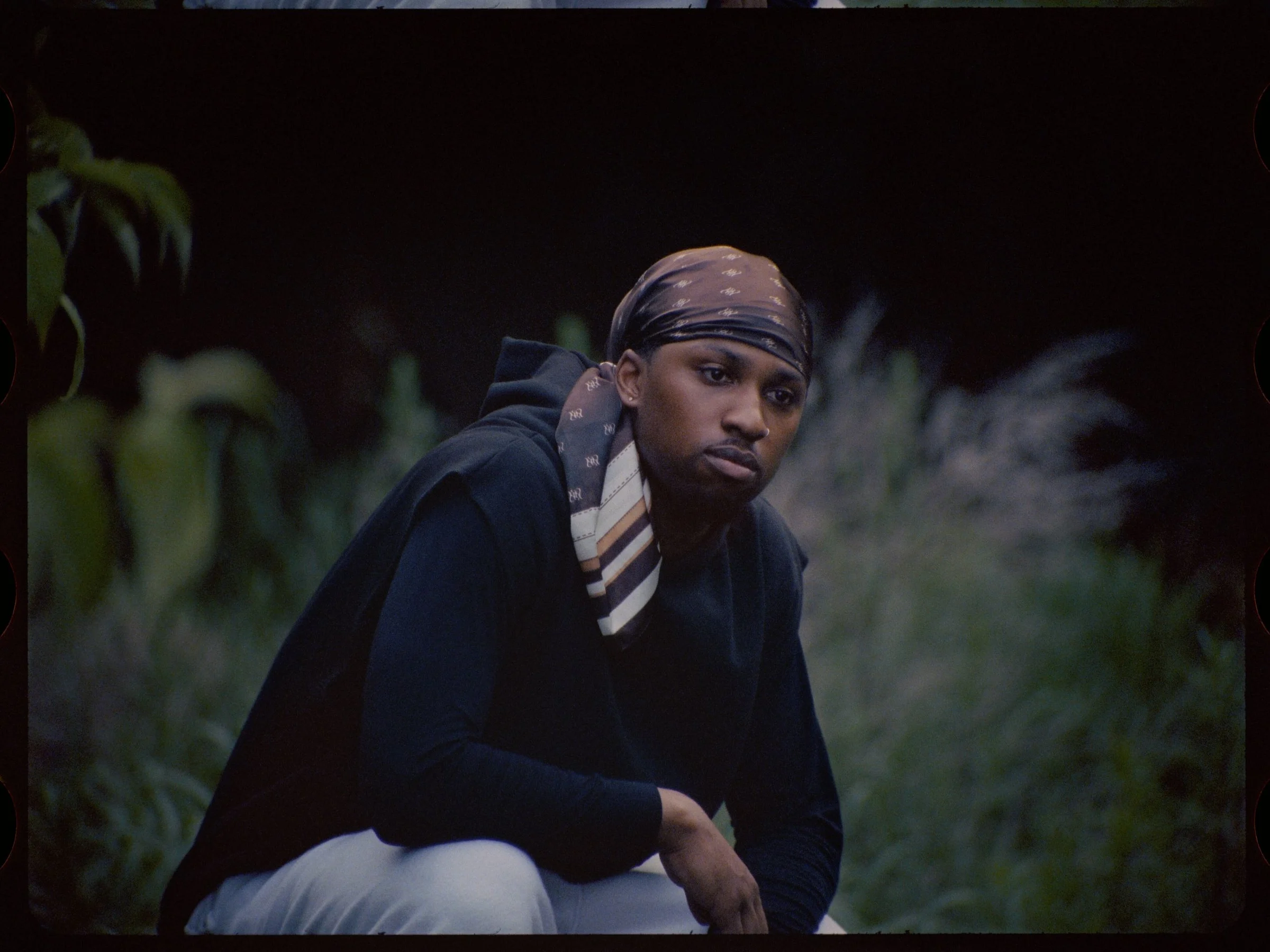 Destin Morisset in a dark hoodie and bandana crouching in tall grass at dusk