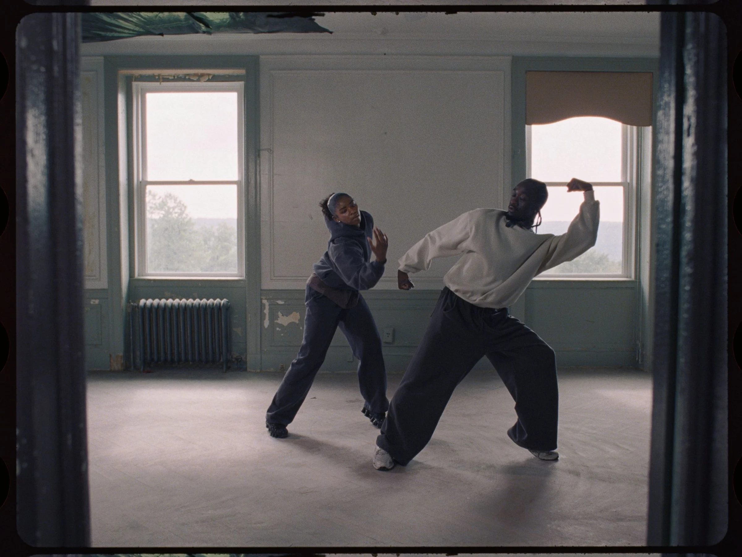 Two dancers mid-motion in an empty room with tall windows