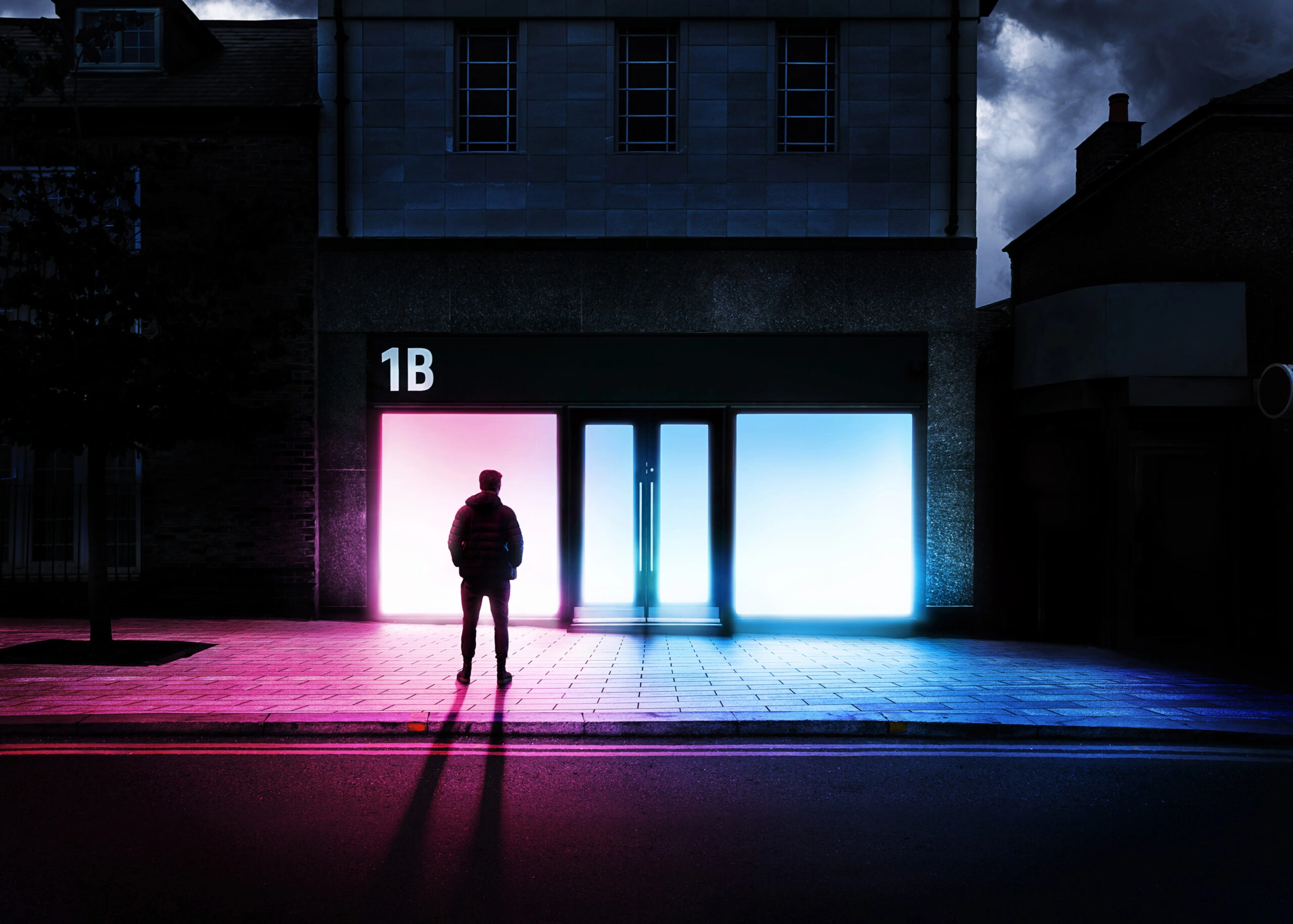 The 1B Window Gallery at night, its windows glowing pink and blue, with a figure standing on the pavement outside