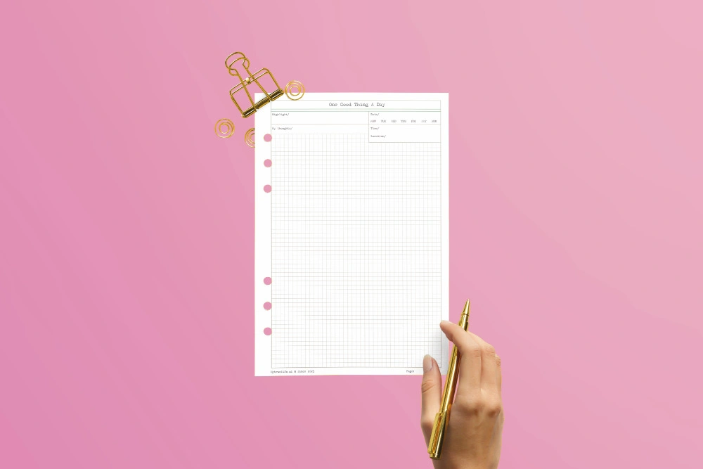 One Good Thing A Day A5 ring binder insert held up with a gold pen and gold binder clips on a pink background